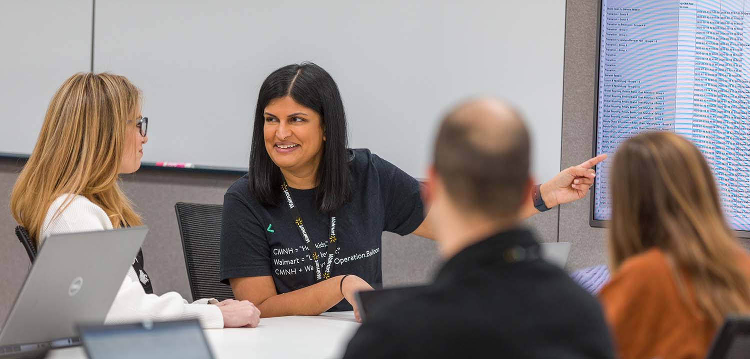 Photo of a tech team gathering and collaborating in a conference room.  A female is discussing a topic with her 3 colleagues.