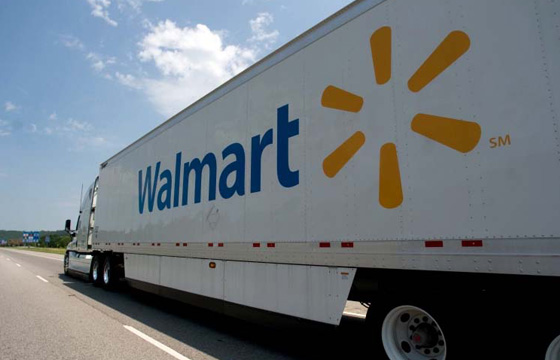 Photo of Walmart semi-truck and trailer driving on a freeway away from the camera with the trailer decorated with the Walmart branding in blue accompanied by the yellow Walmart Spark.