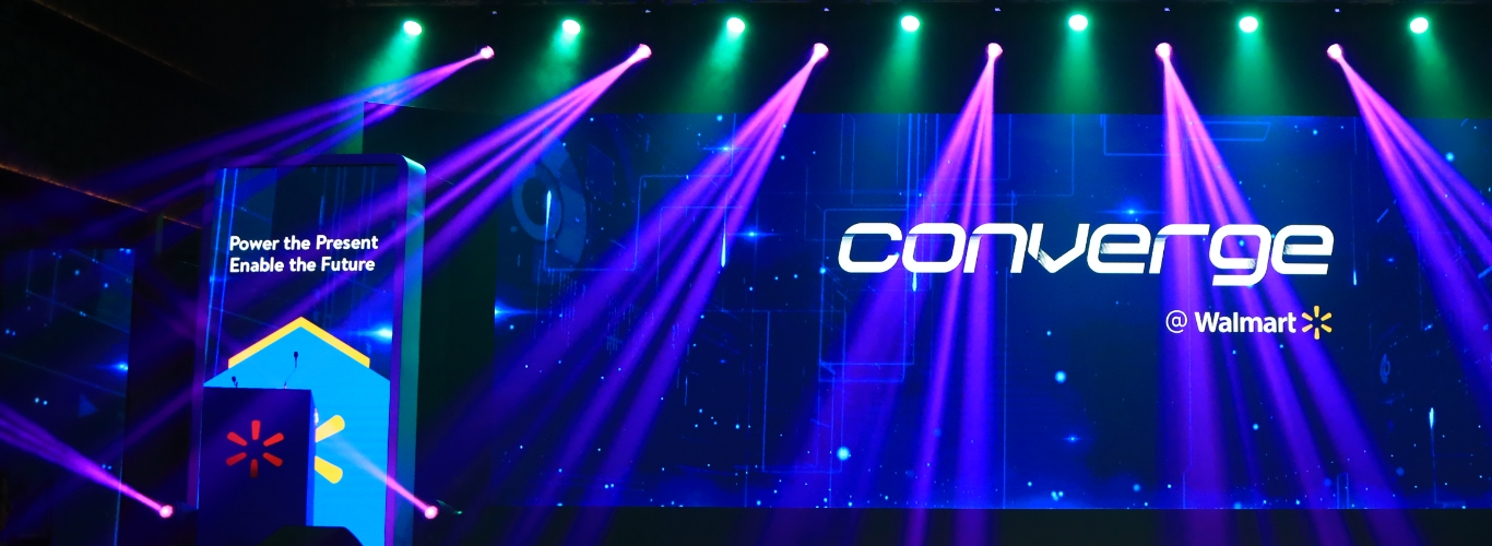 The stage setup for Converge @ Walmart, with purple light beams. In the center of the stage is a large screen which says ‘Converge @ Walmart’. On the left of the stage is a screen that says ‘Power the Present, Enable the Future’.
