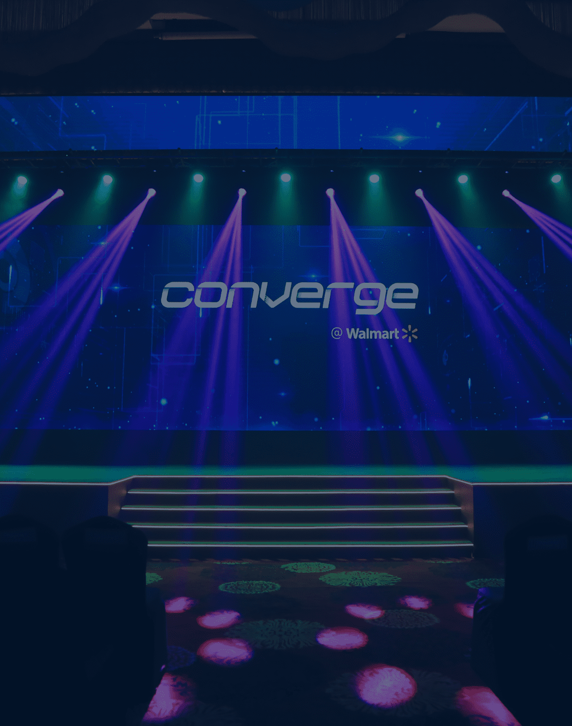The stage setup for Converge @ Walmart, with purple light beams. In the center of the stage is a large screen which says ‘Converge @ Walmart’. On the left of the stage is a screen that says ‘Power the Present, Enable the Future’.