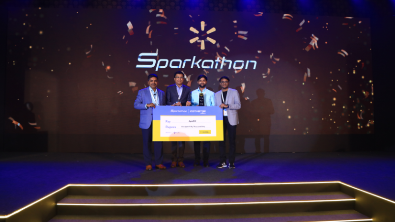 Balu Chaturvedula, SVP and Country Head, Walmart Global Tech, and Hari Vasudev, EVP, Global Tech Platform, Walmart Global Tech, stand together on stage at Converge @ Walmart with two winners of Sparkathon holding a cheque that was awarded to them.