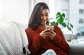 Image of a woman sitting inside a room on a couch. She is smiling and looking at the screen of a smartphone.