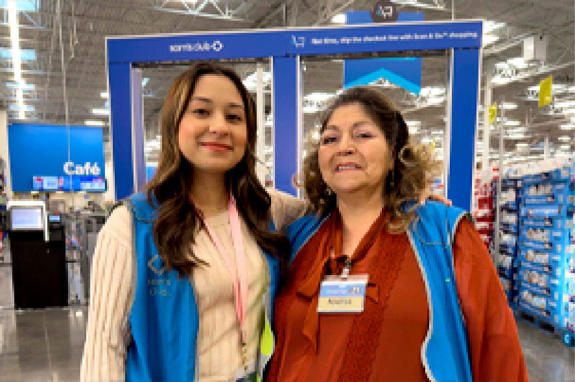 The image shows two associates smiling and standing in front of the blue exit archways inside a Sam’s Club store.