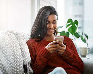 Image of a woman sitting inside a room on a couch. She is smiling and looking at the screen of a smartphone.