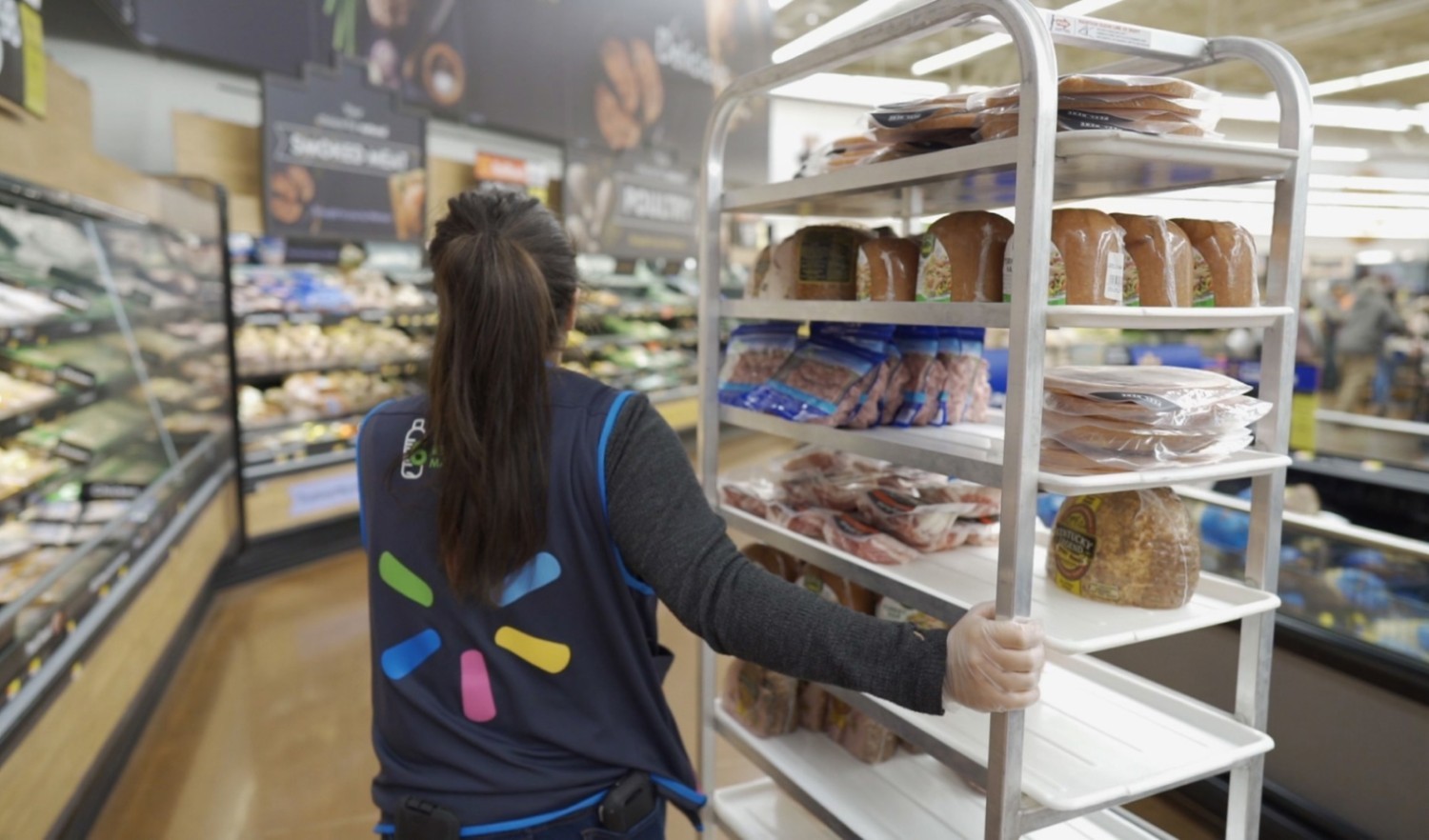A female employee wearing a vest with Walmart Spark logo on the back pulls a tall bakery trolley with food through a bakery section at a supermarket